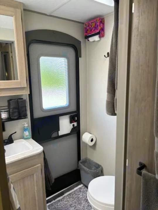 This functional RV rental bathroom includes a sink, toilet, and entry door. Towels, a trash can, and a tissue box are visible.. Forest River Aurora Aurora 2024
