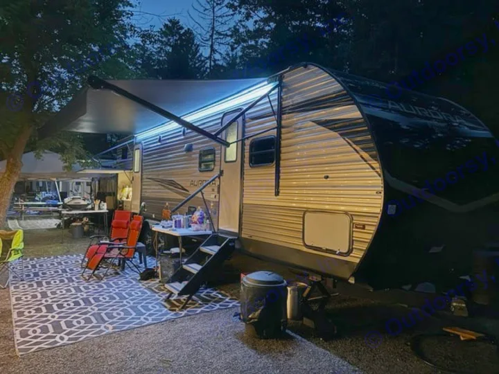 Cozy RV rental campsite at night with illuminated awning, outdoor rug, and seating setup, perfect for evening relaxation.. Forest River Aurora Aurora 2024