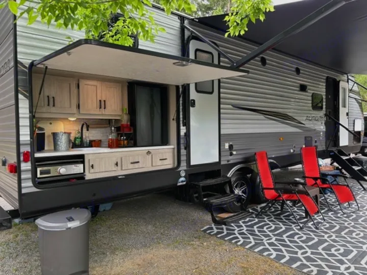 Relaxing rv rental setup with an outdoor kitchen, extended awning, and red chairs for comfortable outdoor living.. Forest River Aurora Aurora 2024