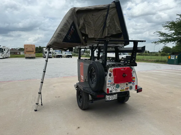 An equipped camping trailer with a 23ZERO rooftop tent displayed at an RV rental lot, surrounded by RVs and rental vans.. taxa Woolly Bear 2017