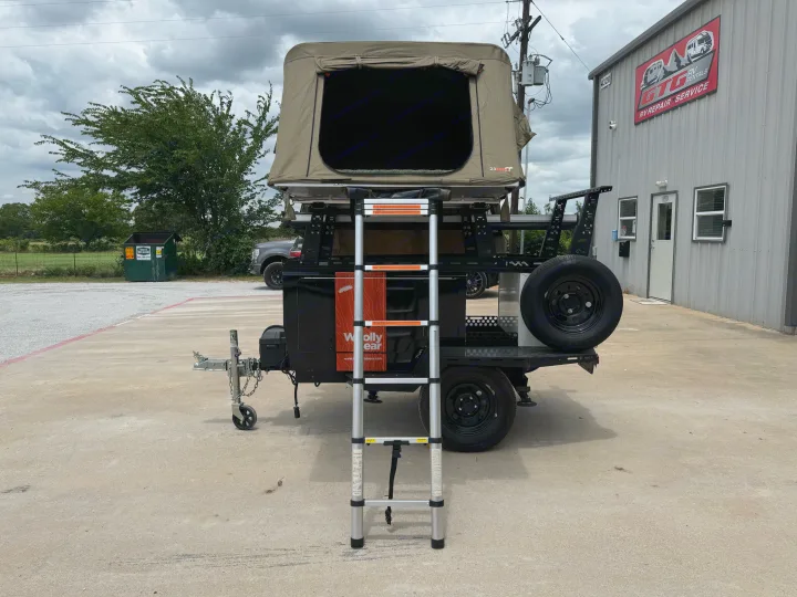 A camper trailer with an open rooftop tent and ladder, parked outside an RV rental and repair service building.. taxa Woolly Bear 2017