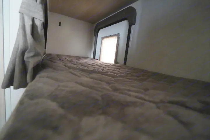Low angle shot of a dark quilted bunk bed mattress inside an RV rental, with a bright window and curtain at the end.. Forest River Rockwood 2021