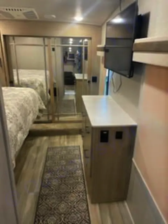 This RV rental bedroom features mirrored closet doors, patterned bedding, a dresser with a TV, and a runner rug on light flooring.. Forest River Rockwood Ultra Lite 2022
