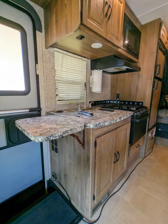 Compact RV rental kitchen features wood cabinets, speckled counter, sink, stove, microwave, and an extendable side table.. Forest River Coachmen Prism 2018