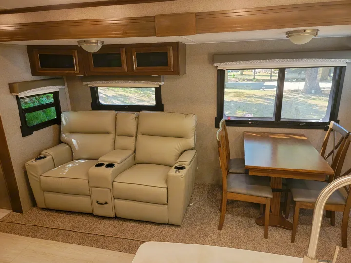 Cozy RV rental interior with a light beige reclining sofa, wooden dining table, and views through large windows.. Forest River Rockwood Signature 2020