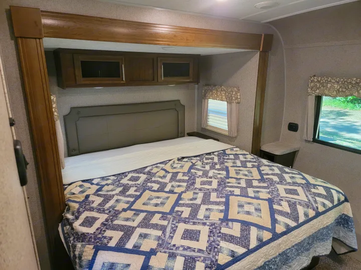 RV rental bedroom: Comfy bed with blue quilt, padded headboard, wooden cabinets, and window.. Forest River Rockwood Signature 2020