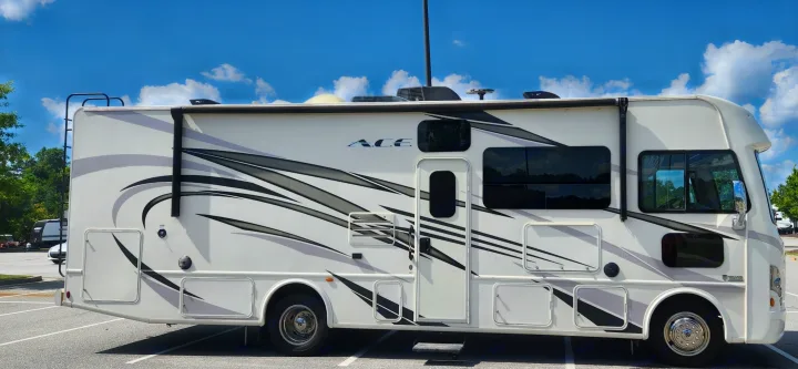 Side view of a white "ACE" RV rental, a motorhome with black and gray graphics, parked on asphalt under a clear sky.. Thor A.C.E. 2018