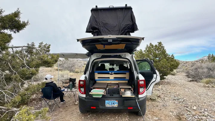 White SUV RV rental setup for car camping, featuring a rooftop tent & rear camp kitchen. Desert scene with person relaxing under awning.. Ford Bronco Sport 2022