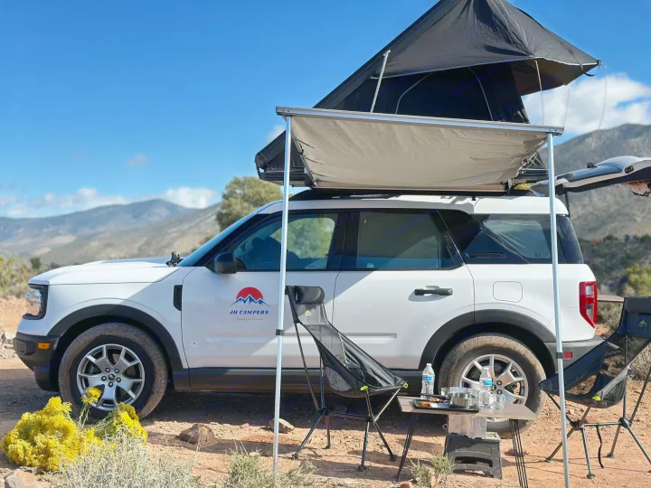 A white SUV, an RV rental, with a rooftop tent & awning set up for camping in mountains.. Ford Bronco Sport 2022