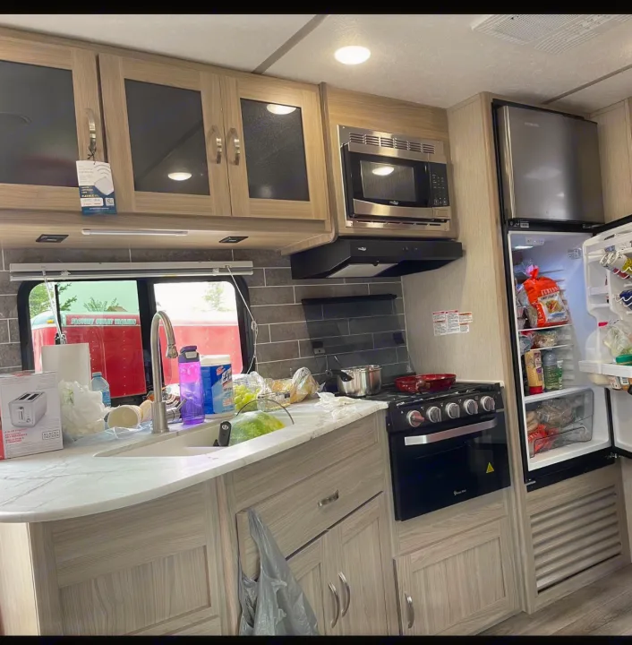 Inside an RV rental, this kitchen features a light wood interior with a sink, microwave, stove, and a full, open refrigerator.. Coachmen fet257bhs-320 2021