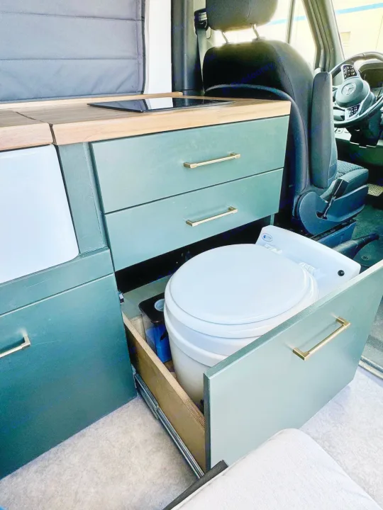 Inside an RV rental: dark green cabinets with gold handles, a kitchen counter with a cooktop, and a hidden pull-out toilet.. Mercedes-Benz Sprinter 2021