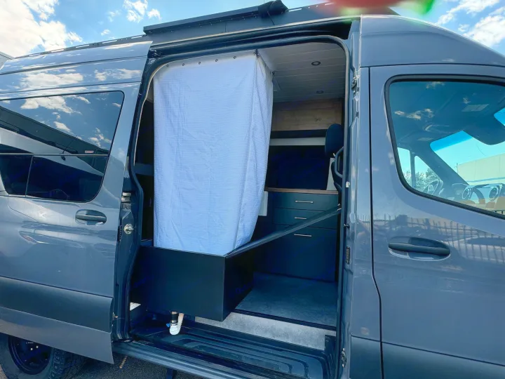 Step inside this RV rental camper van! The side door is open, revealing a clever shower setup with a white curtain & dark cabinetry.. Mercedes-Benz Sprinter 2021