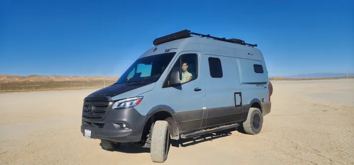 A gray Winnebago Revel RV rental, with a person in the driver's seat, sits on a wide, flat desert plain under blue skies.. Winnebago Revel 2022