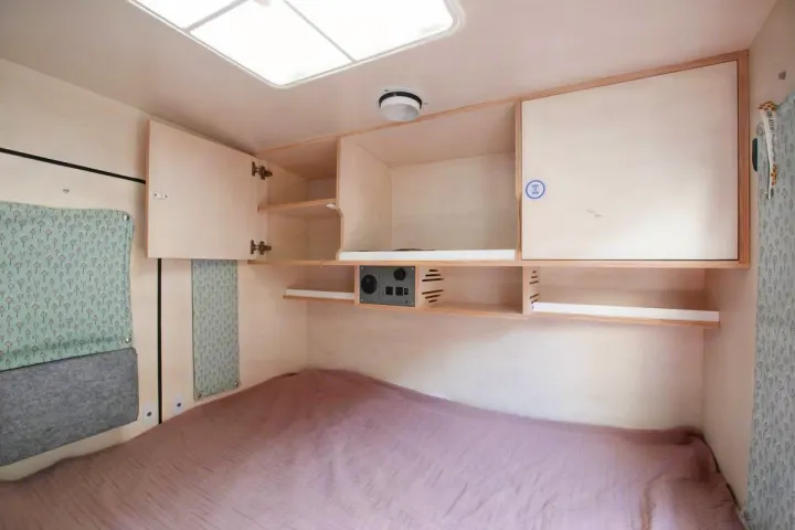 Inside a bright RV rental: a neat bed, custom overhead storage, and a tech panel on wood walls.. Droplet Teardrop 2020