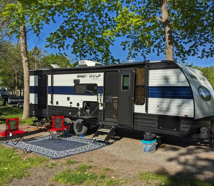 A grey and white rv rental is parked at a sunny campsite with red chairs and an outdoor mat under tall trees.. Forest River Cherokee Grey Wolf 23DBH 2024