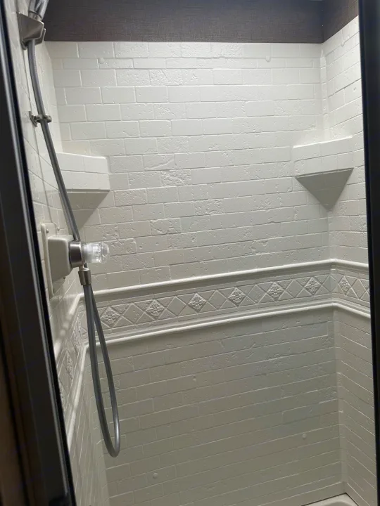 Compact RV rental shower with white textured walls, a handheld shower, and built-in corner shelves.. Thor Hurricane 2017