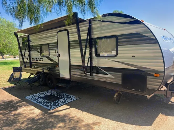 Spacious RV rental, an EVO travel trailer, parked under trees with an extended awning, outdoor mat, and chairs.. Forest River EVO Select 268 BH 2022