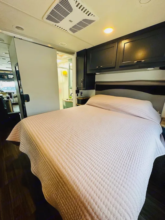 Inside an RV rental: a cozy bedroom with quilted bed, overhead cabinets, AC, and a sliding door to the bathroom.. Thor Motor Coach A.C.E 2018