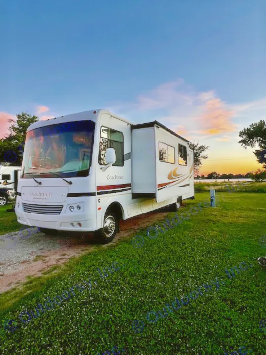 A white Coachmen Mirada RV motorhome with extended slide-out, perfect for an RV rental, parked on grass & gravel at sunset.. Forest River Coachmen Mirada 32 DSF 2011