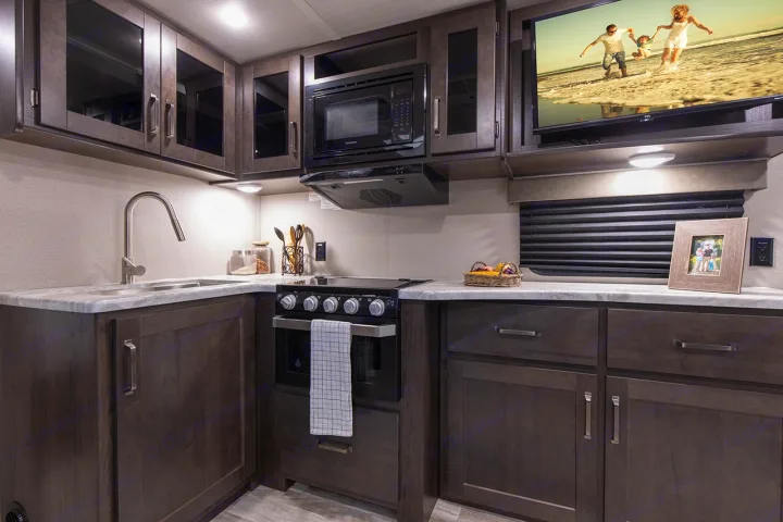 Modern RV rental kitchen with sink, stove, microwave, and TV. Ample dark wood cabinets and light countertops.. Grand Design Transcend Xplor 221RB 2023