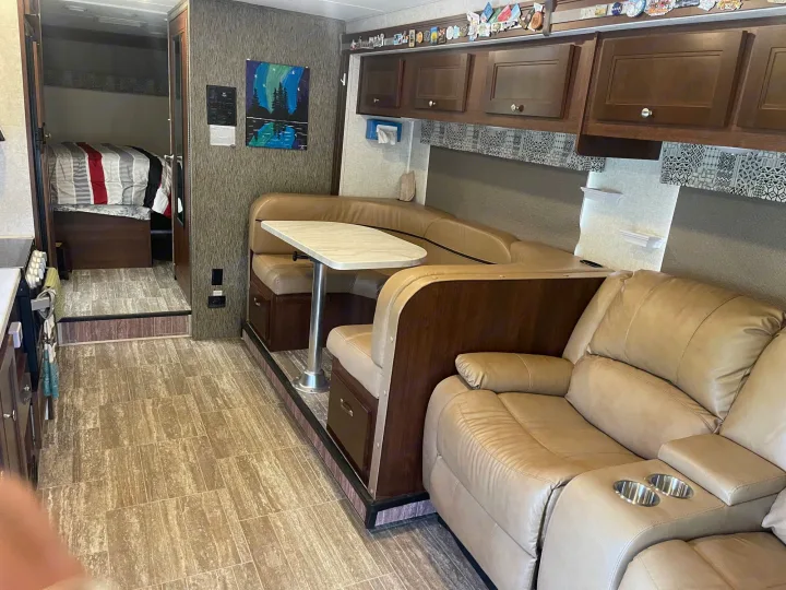 RV rental interior showing a dining booth, comfy recliners, and a bedroom area with striped bedding.. Forest River Sunseeker 2018