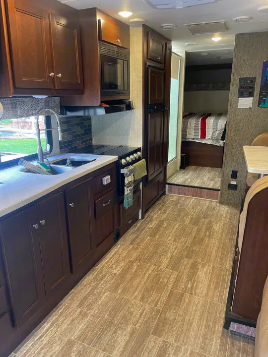 RV rental interior featuring a full kitchen with dark wood cabinets, sink, stove, microwave, and fridge, plus a cozy bedroom area.. Forest River Sunseeker 2018