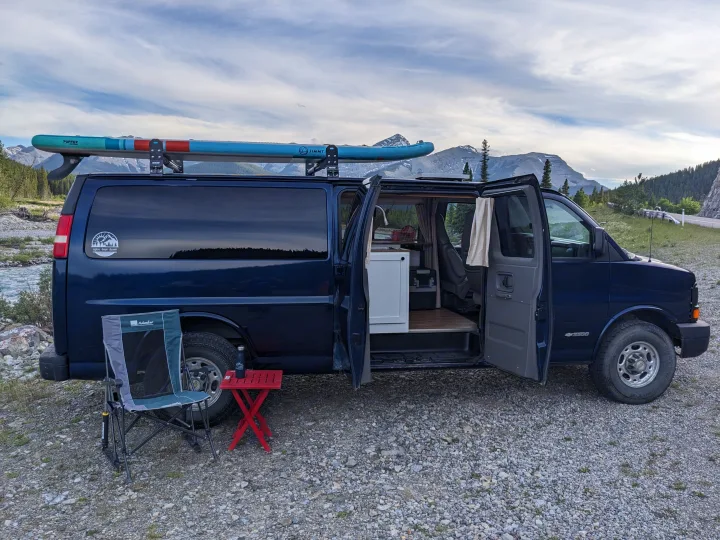 This dark blue camper van, available as an RV rental, is set up for adventure by a river, with mountains, a paddleboard, and outdoor seating.. Chevrolet Express 2003