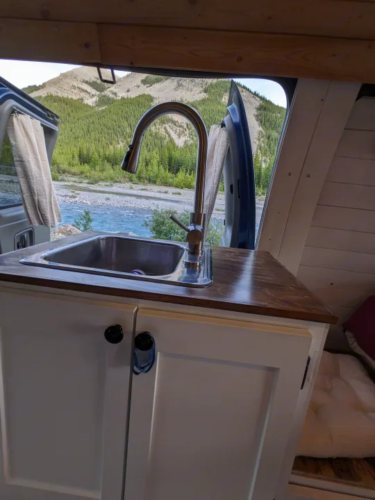 RV rental's functional kitchen featuring a sink and faucet, with breathtaking views of mountains and a river through the open door.. Chevrolet Express 2003