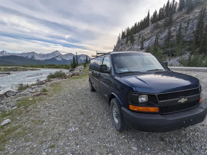 Blue RV rental van on gravel by a flowing mountain river, with majestic snow-capped peaks and forested slopes.. Chevrolet Express 2003