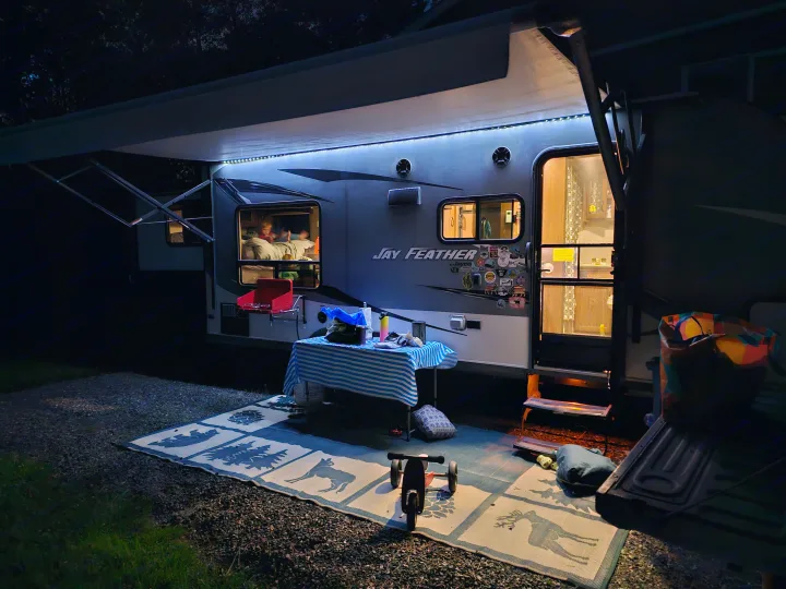 Cozy night at a campsite with an illuminated RV rental. People are visible inside, while an outdoor table and mat are set up.. Jayco X213 2016