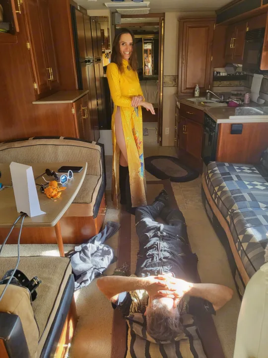 Smiling woman in a yellow dress in an RV rental, beside a man relaxing on the sunlit floor. Cozy RV interior with kitchen.. Fleetwood Expedition 2003