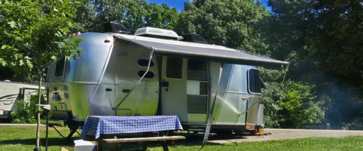 A shiny silver Airstream travel trailer (RV rental) with its awning extended, parked at a sunny campsite with a picnic table.. Airstream Caravel 2022