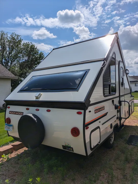 White A-frame camper, great for an RV rental, parked on gravel and grass. Sun glares off its roof under a cloudy blue sky.. Rockwood A122s 2023