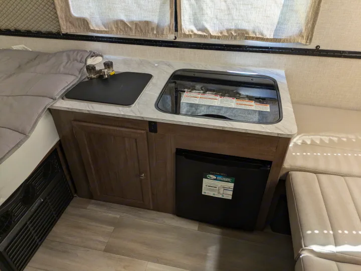RV rental interior featuring a compact kitchen with a covered sink, stovetop, and mini-fridge, adjacent to a bed and bench seating area.. Rockwood A122s 2023
