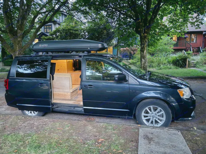 Black minivan converted into an RV rental, showing its custom wood interior and roof box, parked on a street.. Dodge Grand Caravan 2013