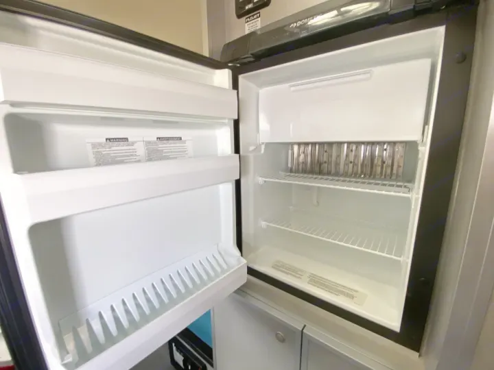 Interior of an open, clean Dometic refrigerator and freezer in an RV rental, showing empty shelves and a top freezer unit.. Helio Helio Trailer 2023
