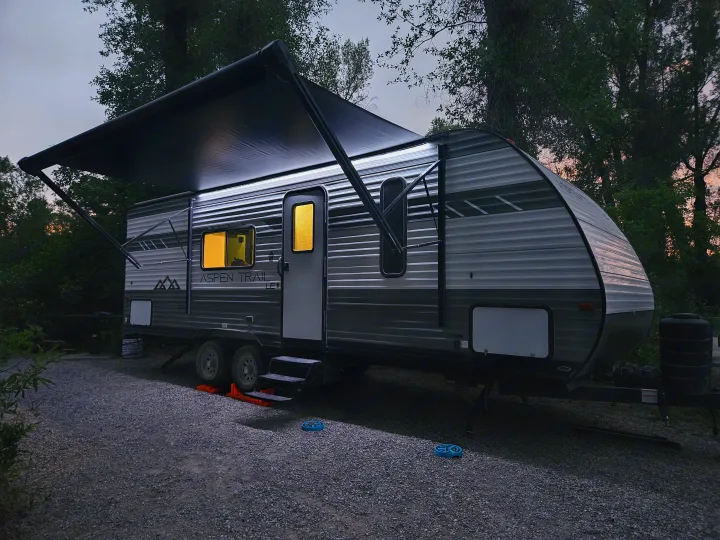 An illuminated RV rental is settled for the evening at a treed gravel campsite, with its awning extended and interior lights glowing.. Dutchmen TT 2024