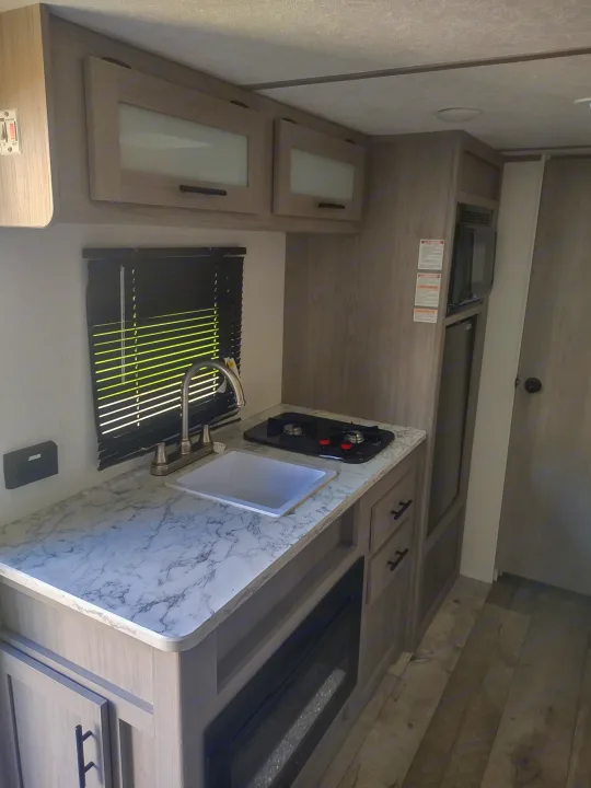 Modern RV rental kitchen featuring a sink, 2-burner stove, microwave, fridge, and a built-in electric fireplace.. Coachmen 17b 2024