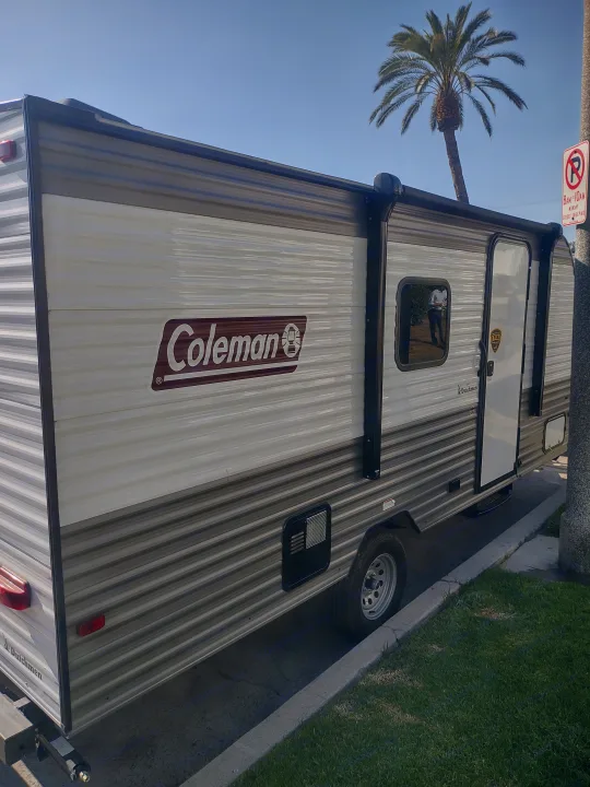 This Coleman travel trailer is a perfect RV rental option. Seen from the side, parked curbside with a visible window, door, and palm tree.. Coachmen 17b 2024