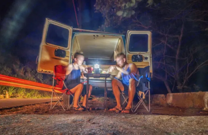 Couple relaxing at night by their open RV rental camper van, illuminated by a lantern, with light trails.. Ford E250 Eagle Camper 2006