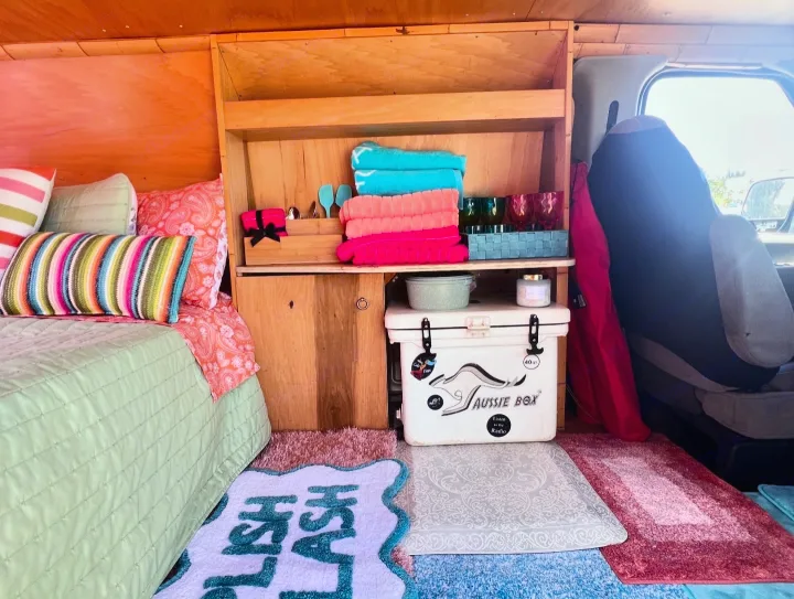 Vibrant RV rental interior: bed, organized shelves with towels/glasses, a cooler, and colorful rugs. Ready for van life adventures.. Ford E250 Eagle Camper 2006