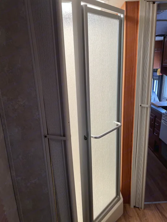 View inside an RV rental showing a compact shower with a textured frosted glass door and an adjacent pleated privacy screen.. Coachmen Freelander 2007