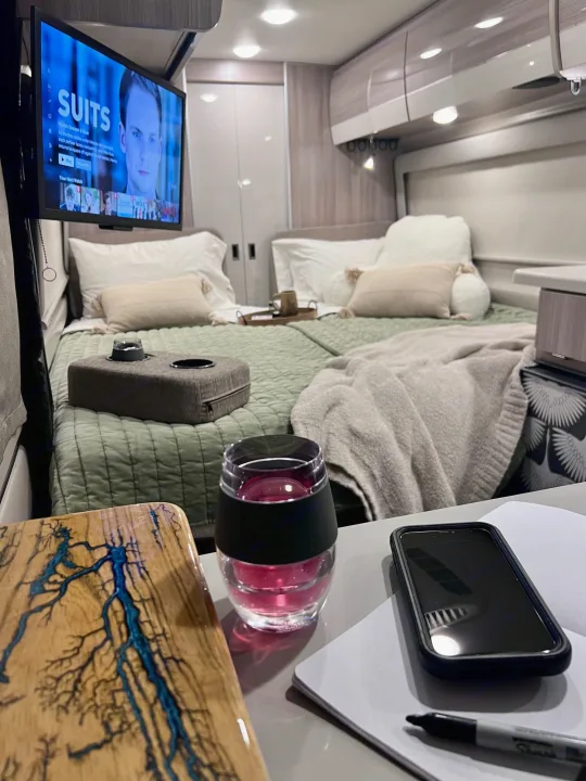 Inside a modern rv rental: a cozy bed with a TV playing "Suits" and a side table holding a drink, phone & notebook.. Thor Sequence 2023