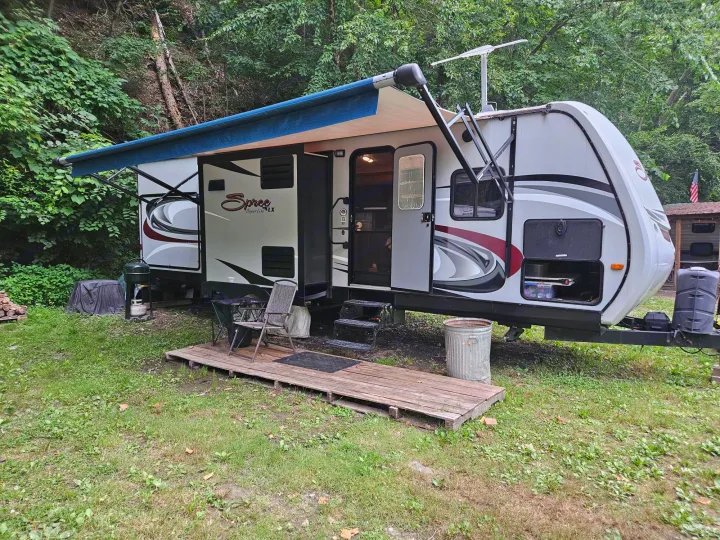 An RV rental travel trailer with an extended awning and outdoor setup, nestled in a green, wooded campsite.. KZ Spree 301RIK 2015
