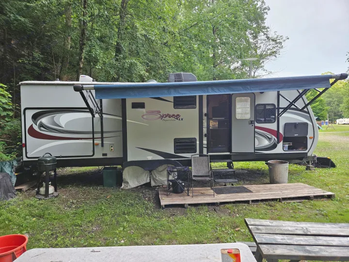 A grey travel trailer RV rental with a blue awning, parked at a wooded campsite, showing outdoor grill and seating.. KZ Spree 301RIK 2015