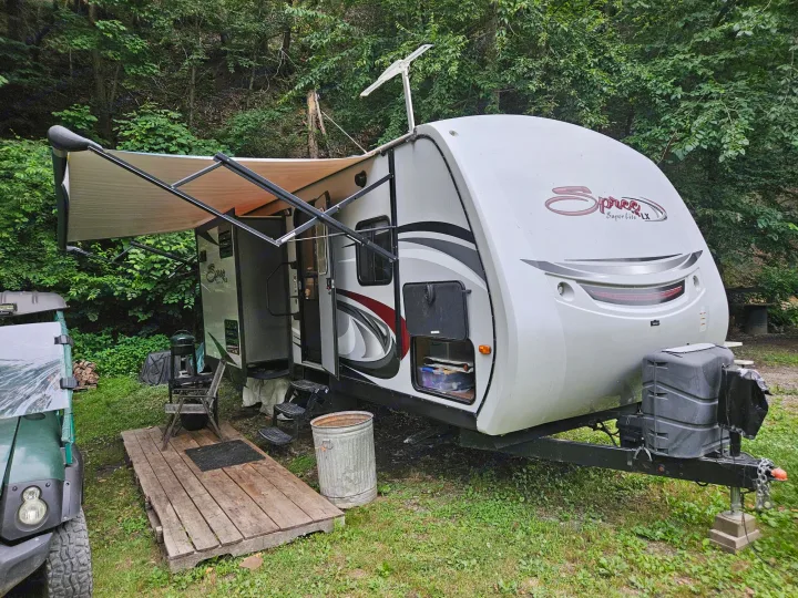 Travel trailer RV rental with awning extended at a green campsite. Features a wooden deck, grill, and UTV.. KZ Spree 301RIK 2015