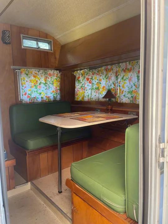 Cozy vintage RV rental dinette featuring green cushions, wood paneling, floral curtains, and a table with a small lamp.. Forest River Forester 1967