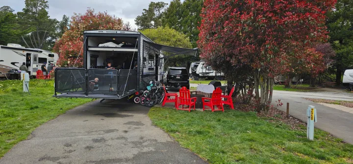 Family enjoying their RV rental, set up with a toy hauler patio, red chairs, and bikes at a scenic campsite.. Keystone Fuzion 2024