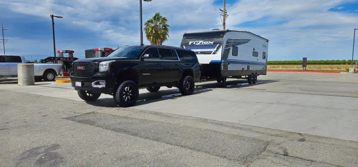 Black GMC Yukon XL towing a Fuzion travel trailer, parked at a gas station. Ideal for your next RV rental trip!. Keystone Fuzion 2024