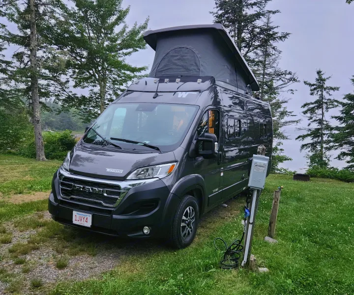 Gray RV rental with pop-top extended at a scenic campsite, hooked up to power, overlooking a serene body of water.. Winnebago Solis 2024